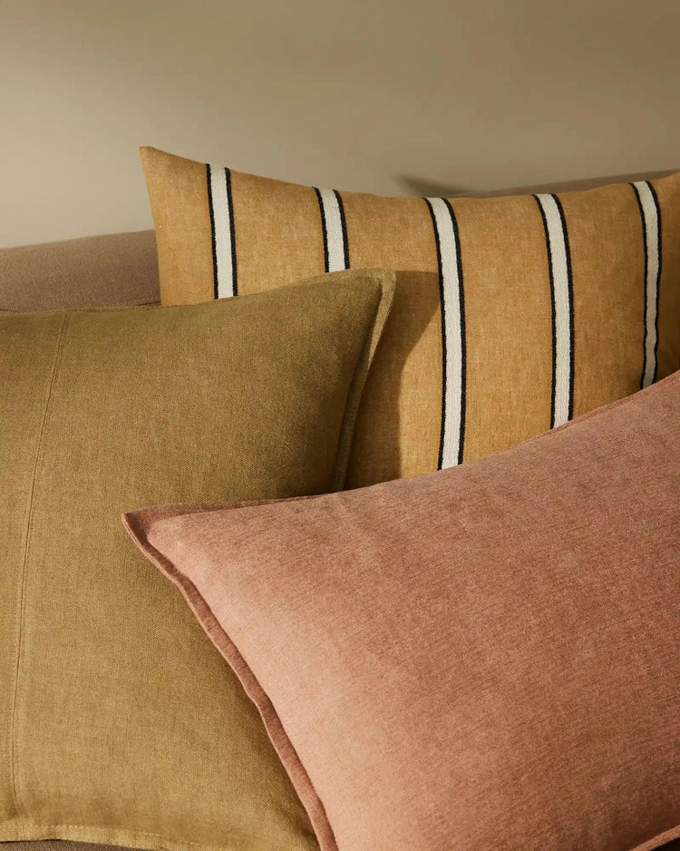 Warm earth-tone linen cushions layered on a neutral sofa, featuring terracotta and ochre textures with black and cream striped details