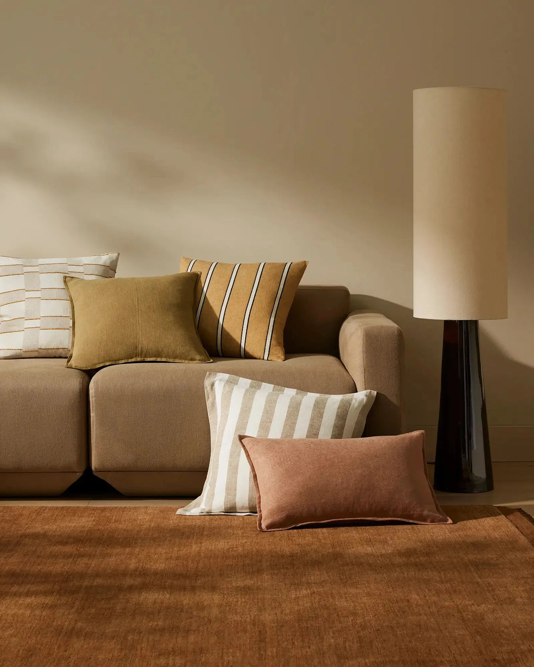 Warm earthy living room with layered Fiore and Vinnie cushions in terracotta, ochre and cream stripes on taupe linen sofa, ochre rug and natural light casting soft shadows on walls