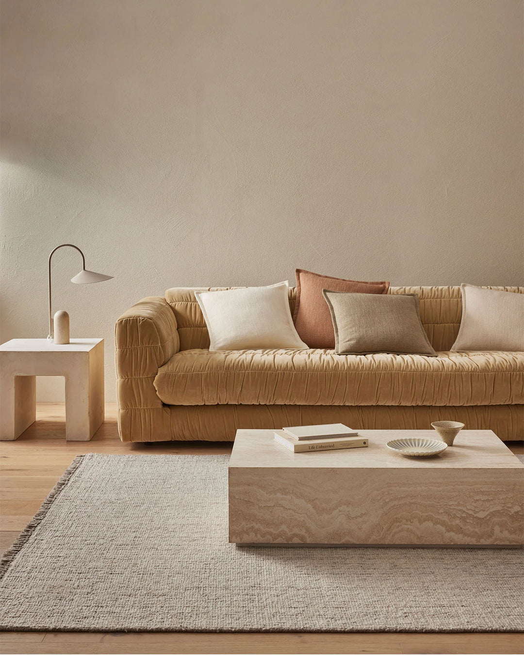 Warm neutral living room with ochre quilted sofa, cream and terracotta cushions, layered natural fiber rug, and minimalist wood furniture in calming earth-tone palette