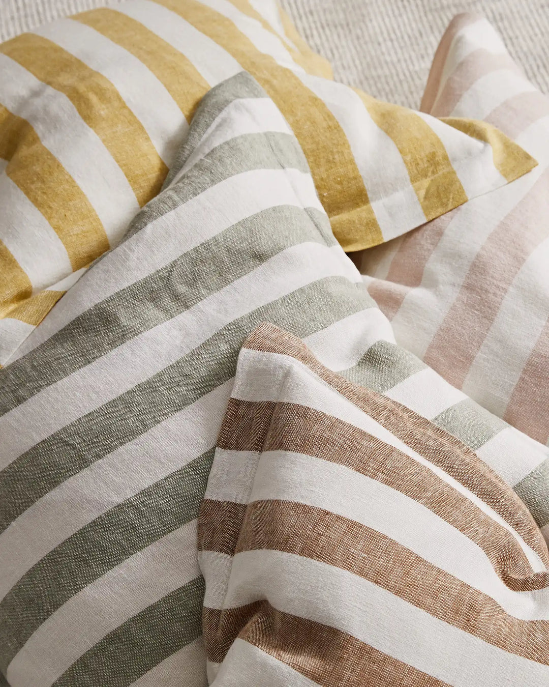 Layered striped linen blend cushions in warm neutral tones—gold, sage, taupe and blush—arranged in soft folds showcasing textured weave and contemporary stripe pattern