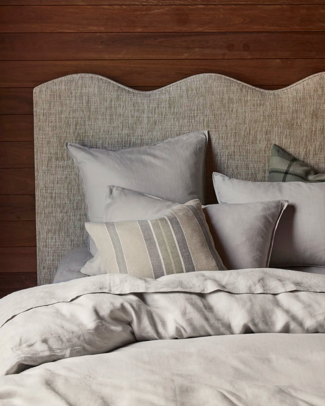 Neutral bedroom styling with layered linen bedding in soft greys and cream tones, featuring textured pillows and striped cushion against a curved upholstered headboard