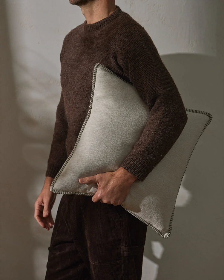 Warm-toned lifestyle scene: man holding Alba cushion in bone with contrast whipstitched edge, layered with chocolate knit sweater and charcoal corduroy against neutral wall backdrop