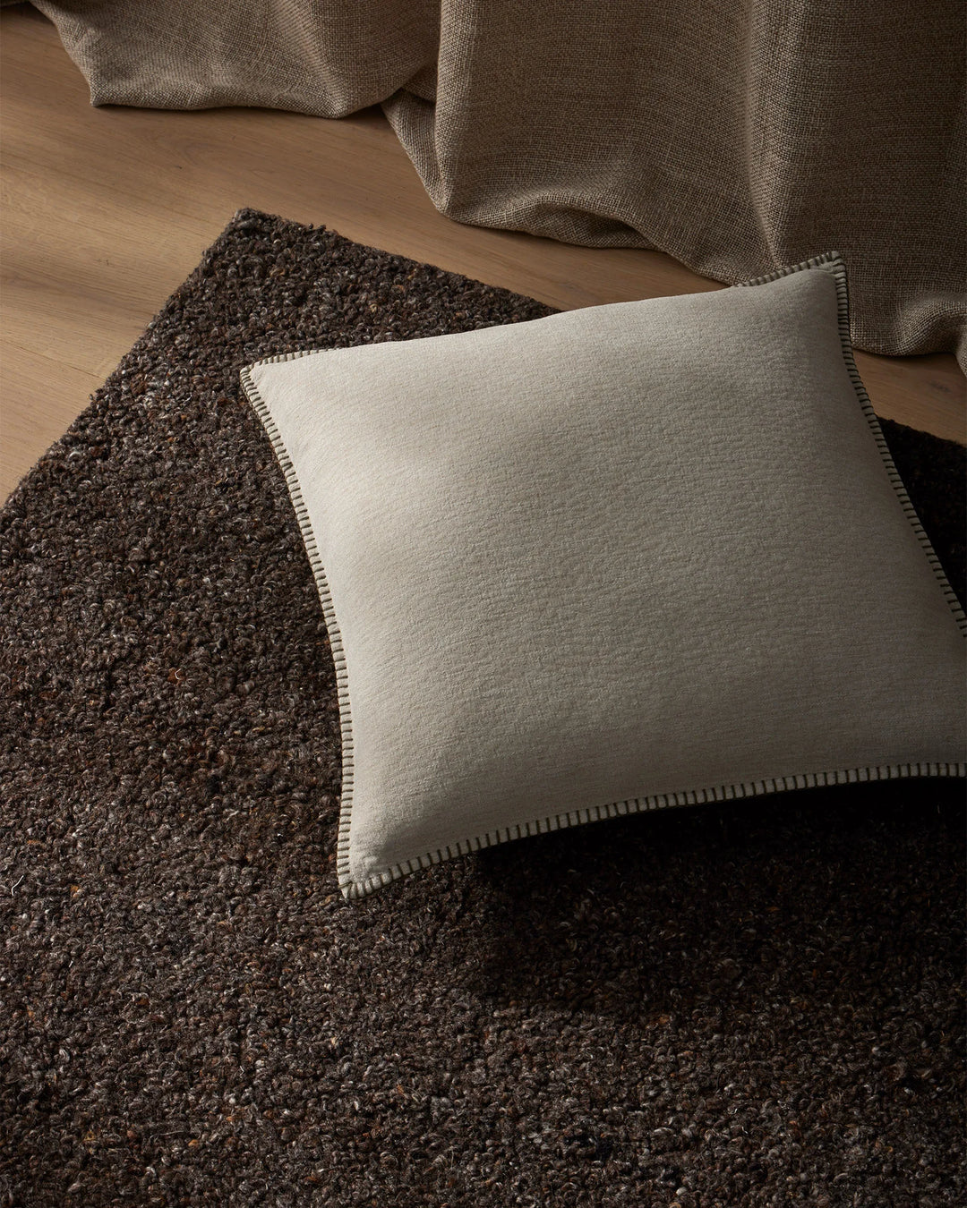 Neutral earthy living room styled with charcoal wool-blend rug, cream linen cushion with whipstitching, and taupe knit throw—warm minimalist palette with texture layering