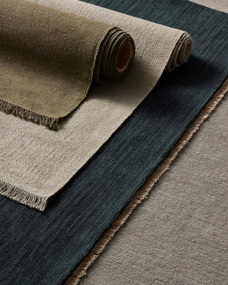 Rolled and layered linen rugs in earthy palette of sage, cream and charcoal, showcasing natural textile weave and fringe detailing against neutral tones