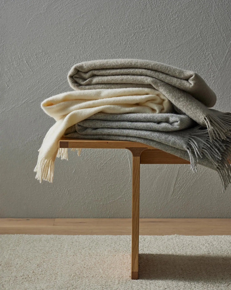 Neutral wool throws in charcoal, cream and grey tones layered on a timber bench against textured plaster wall, showcasing soft monochrome palette and natural fiber textures