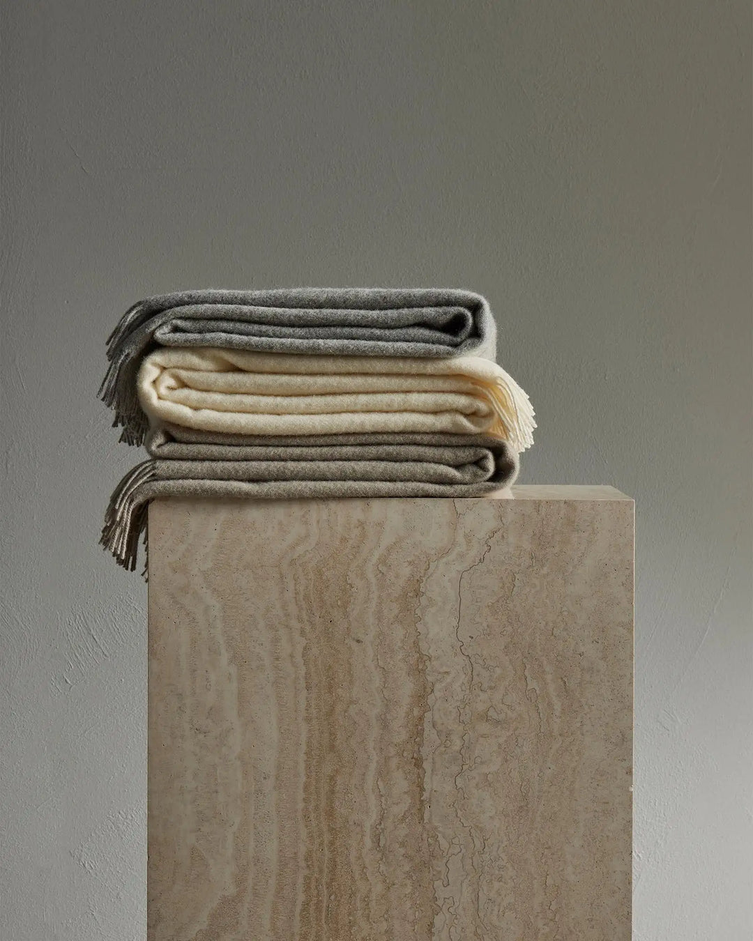 Stacked wool throws in neutral earth tones—ash grey, limestone, and snow—displayed on a textured stone plinth against a soft grey wall, showcasing warm minimalist home styling