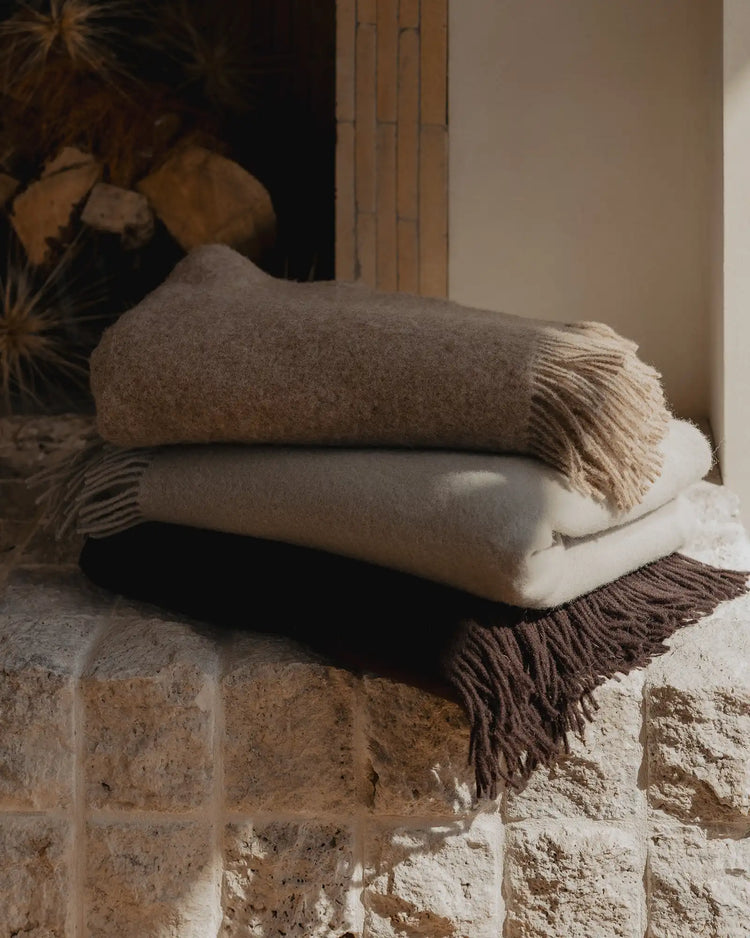 Stacked wool throws in warm earth tones and cream displayed on textured stone, featuring fringed edges and natural fibre textures in a minimalist interior setting