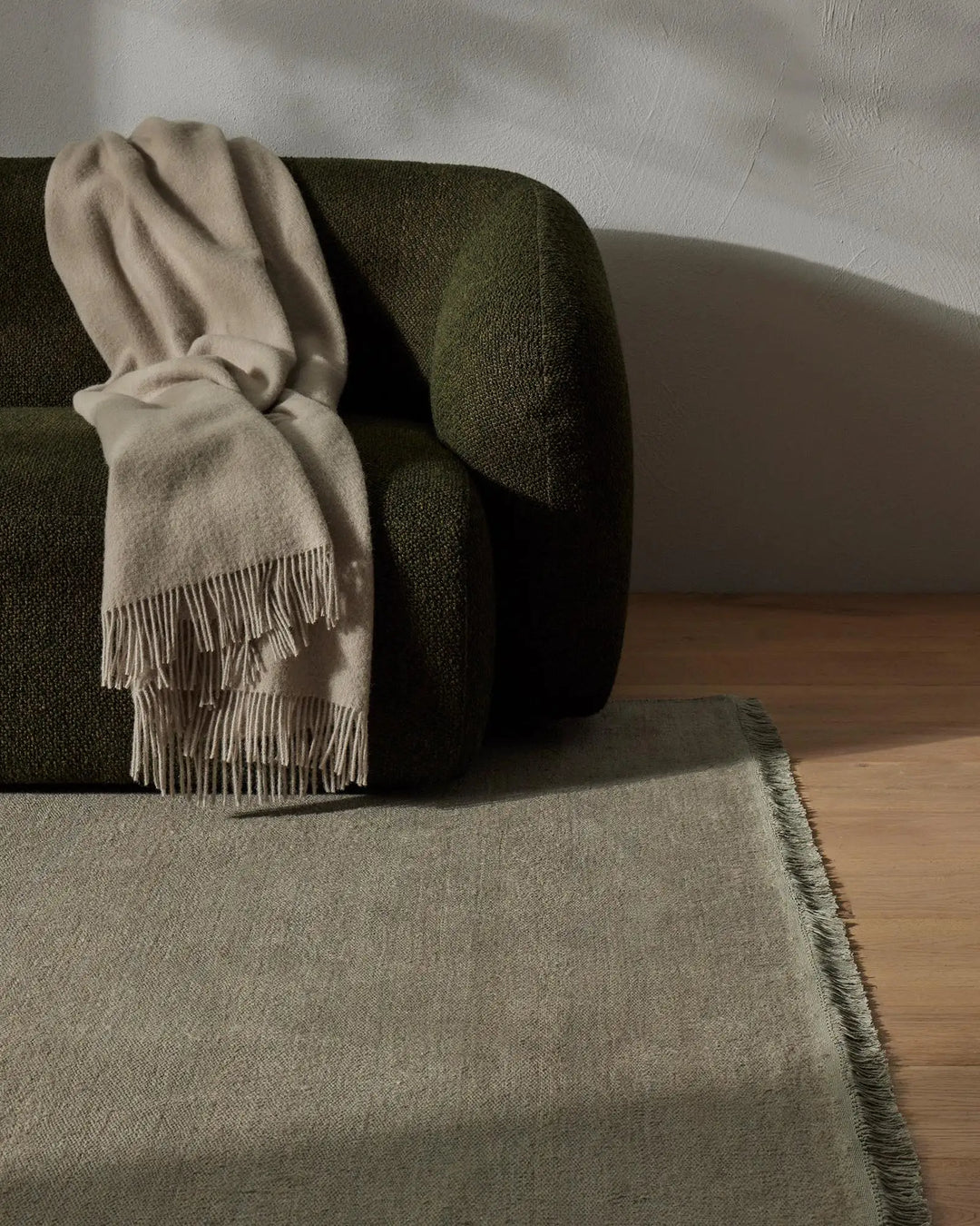 Earthy minimalist living room with olive-green sofa, cream wool throw with fringe detailing, and sage linen rug on warm timber floor against white textured wall