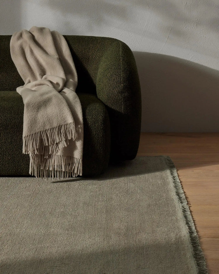 Earthy minimalist living room with olive-green sofa, cream wool throw with fringe detailing, and sage linen rug on warm timber floor against white textured wall