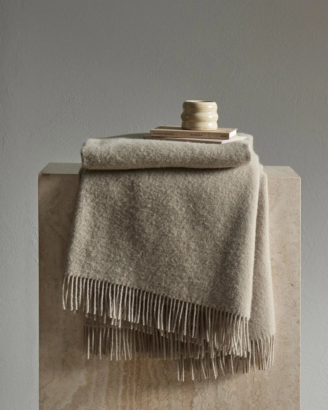 Neutral wool throw with fringe draped over concrete plinth, styled with books and ceramic vessel in minimalist beige toned setting
