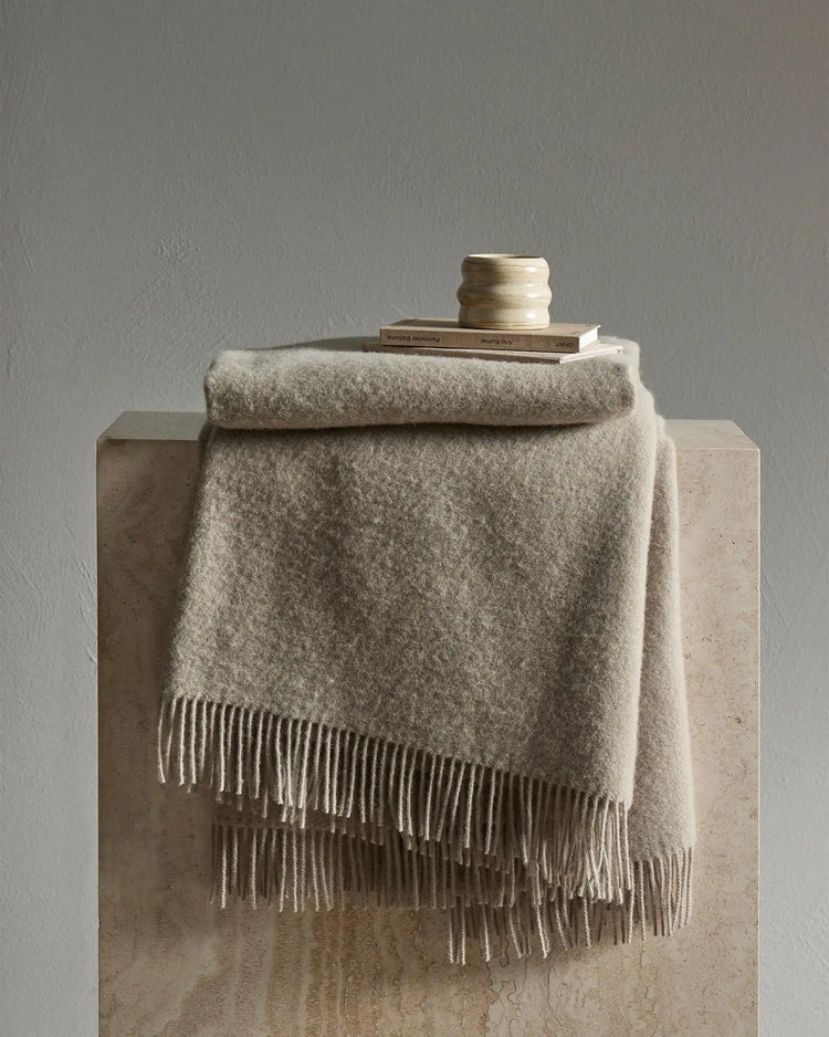 Neutral wool throw with fringe draped over concrete plinth, styled with books and ceramic vessel in minimalist beige toned setting