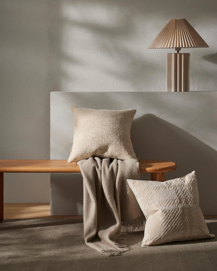 Neutral minimalist interior with textured Nevis throw and Laurent cushions on wooden bench against white wall, warm natural light casting geometric shadows