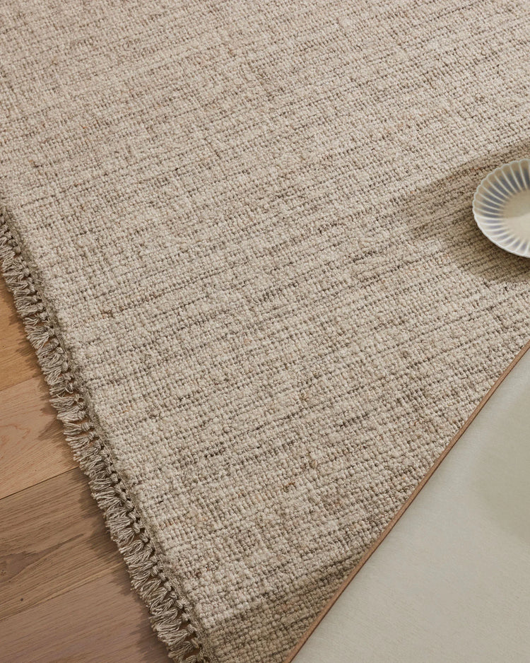 Neutral taupe wool-blend rug with textured weave and fringed edge layered on light timber floor, styled with ceramic dish in minimalist living space
