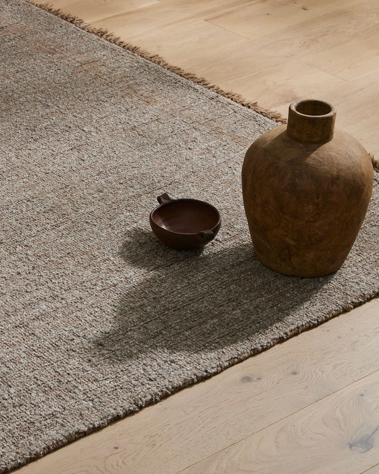 Taupe wool-blend rug with frayed edges anchoring earthy ceramic vessels on light oak flooring, warm neutral palette with organic textures