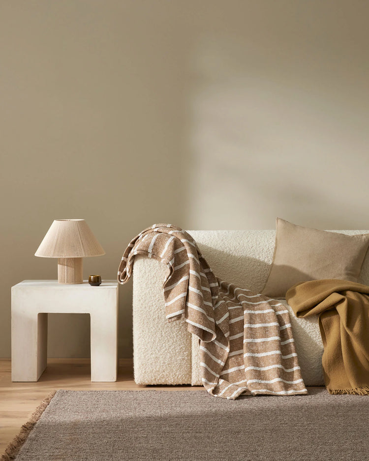 Neutral minimalist living room with cream boucle armchair, taupe and cream striped throw, mustard blanket, white sculptural side table, and natural texture rug in warm earth tones