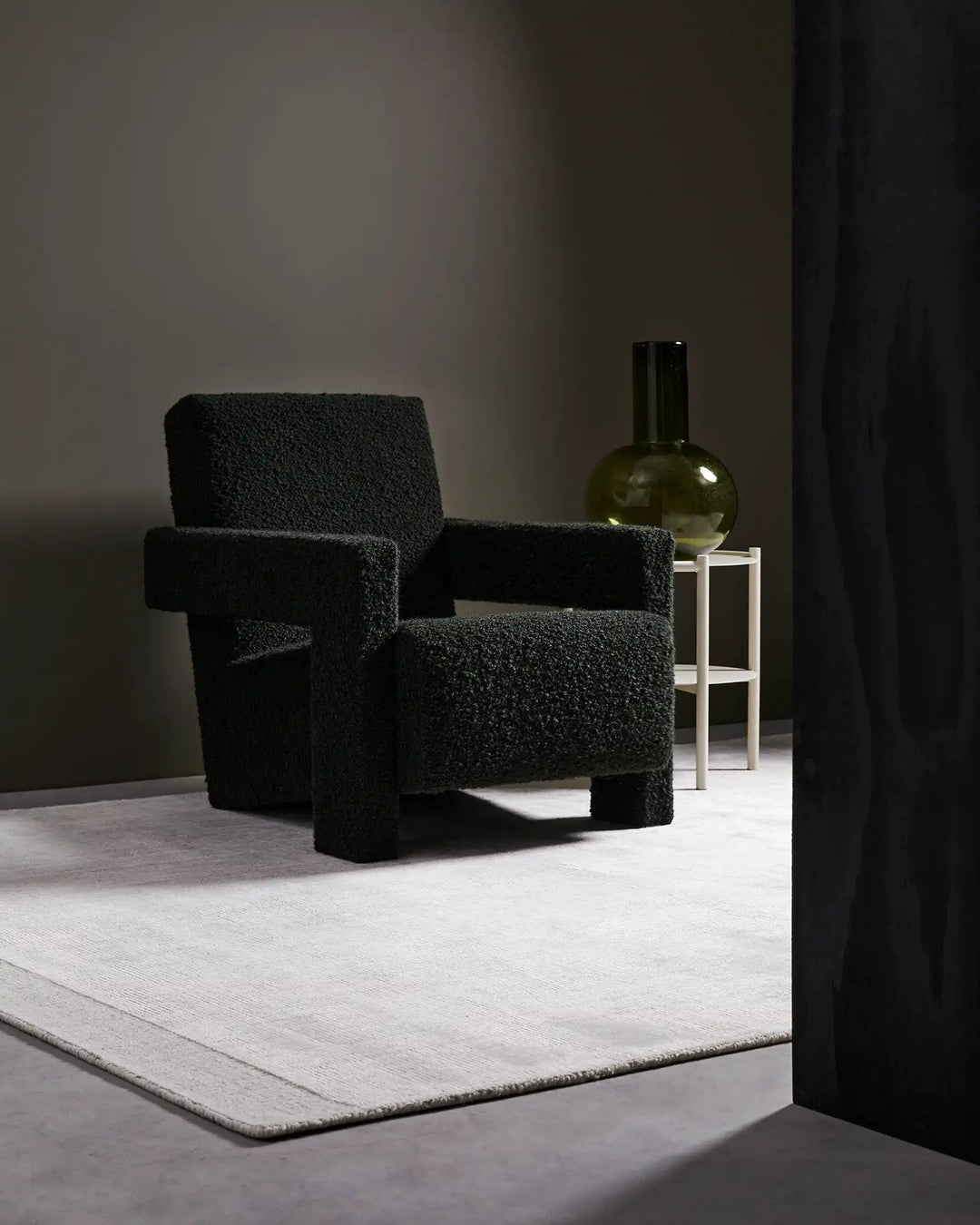 Minimalist living space styled with charcoal boucle chairs on a cream Travertine marble rug, soft neutral tones with olive glass accents and sculptural shadows