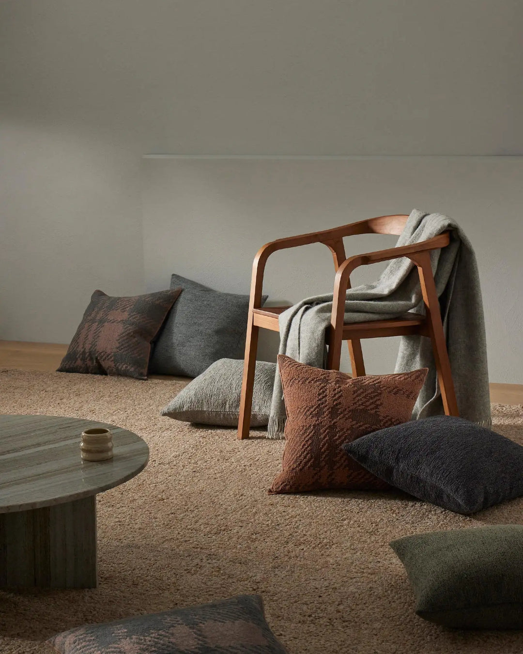 Earthy minimalist living room styled with warm terracotta and charcoal cushions, natural wool rug, and mid-century wood chair draped in cream knit throw