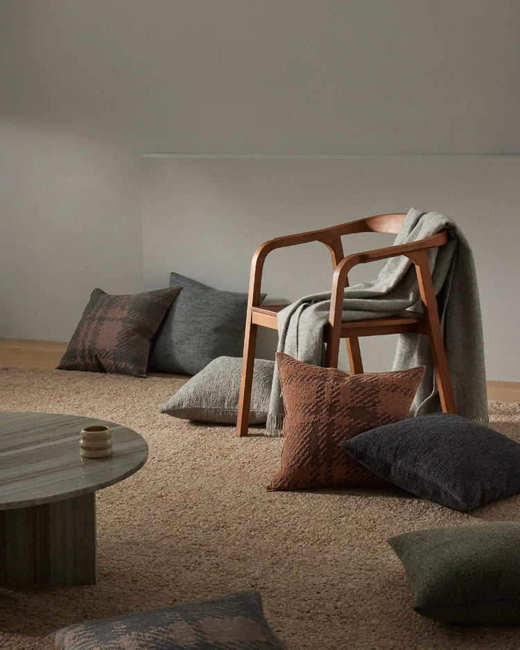 Earthy minimalist living room styled with warm terracotta and charcoal cushions, natural wool rug, and mid-century wood chair draped in cream knit throw