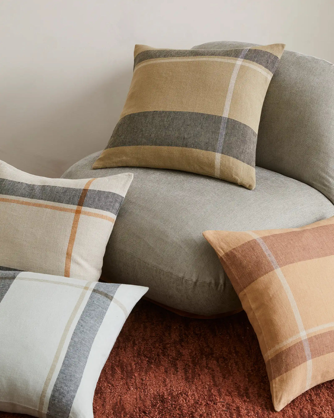 Neutral linen cushions in charcoal, stone and terracotta tones layered on a grey upholstered chair, with warm earth-toned rug below. Minimalist colour blocking design with subtle striping.