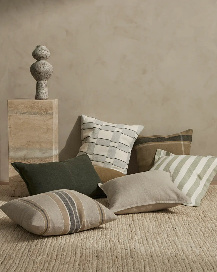 Neutral minimalist styling with layered cushions in cream, olive, and spruce featuring striped and textured linen weaves on natural fibre flooring, sage plaster walls, and travertine surfaces