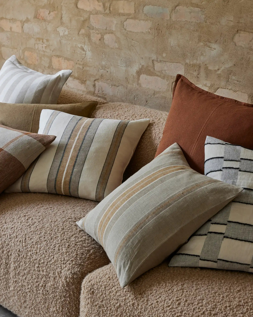 Neutral layered cushions in cream, terracotta and striped linen patterns styled on a textured beige sofa against a distressed brick wall, earthy interior styling