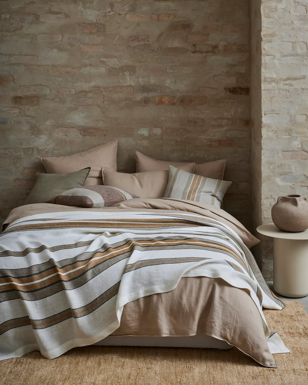 Warm neutral bedroom with whitewashed brick walls, layered in Franco throw with amber and taupe stripes over soft beige bedding, styled with linen cushions and natural textures