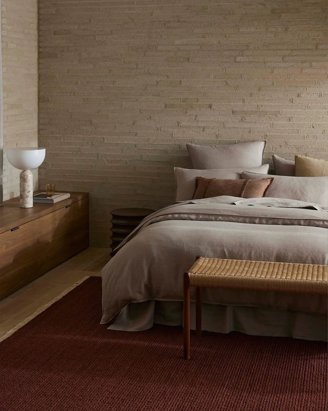 Neutral bedroom styled with warm stone brick, warm timber furniture, and layered bedding in soft taupe with rust and ochre accent pillows. Ravello Fitted Sheet in pebble linen with deep burgundy runner rug creates earthy, minimalist aesthetic.