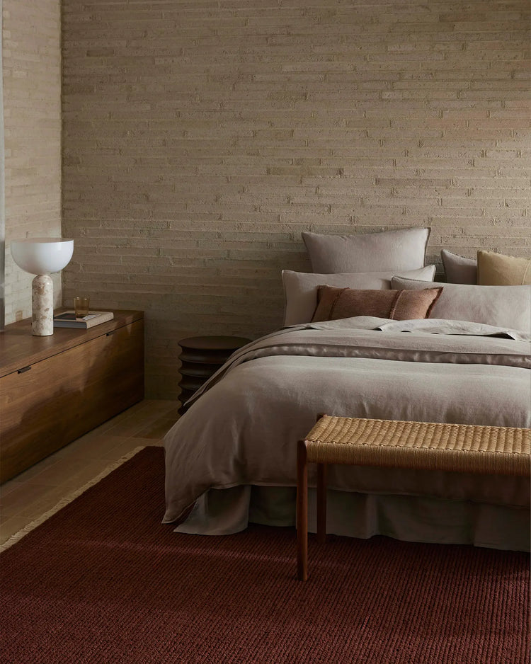 Neutral bedroom styled with warm stone brick, warm timber furniture, and layered bedding in soft taupe with rust and ochre accent pillows. Ravello Fitted Sheet in pebble linen with deep burgundy runner rug creates earthy, minimalist aesthetic.