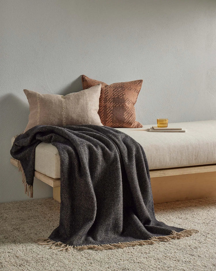 Neutral bedroom scene with layered earth-tone textiles: cream linen cushion with stitch detail, rust jacquard pillow, charcoal herringbone throw with fringe, on pale upholstered bed over warm terracotta rug.