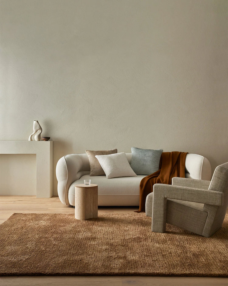 Neutral living room with cream rounded sofa, warm terracotta throw, and layered Tempo cushions in oatmeal and pearl tones against textured jute rug and soft greige walls