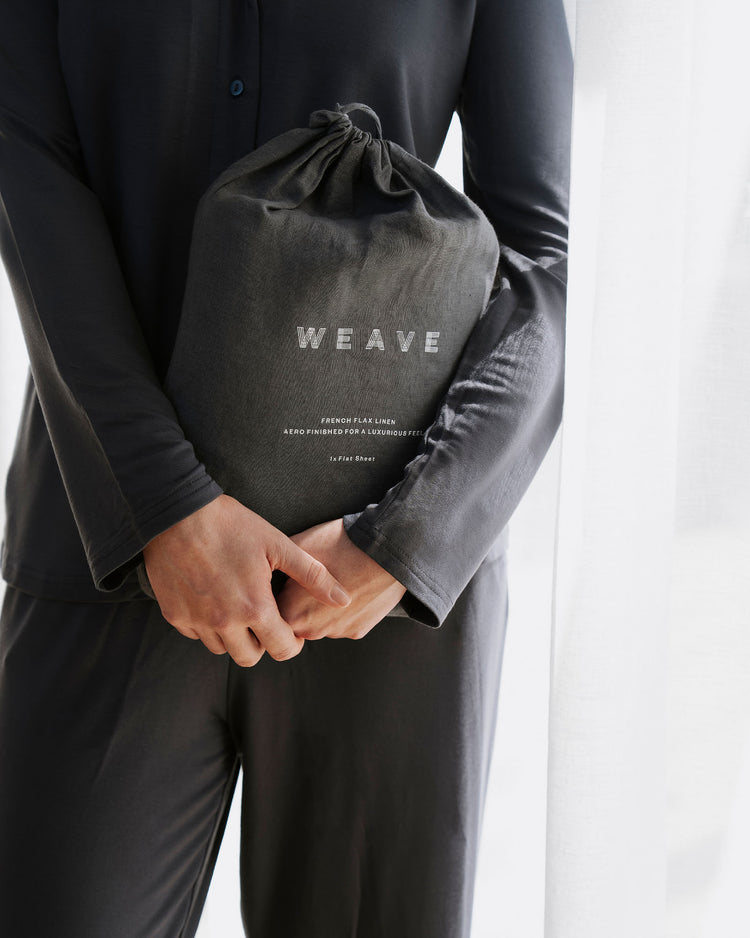 Charcoal linen bedding set in branded drawstring pouch, held against dark minimalist styling, showcasing soft European linen texture and monochromatic luxury aesthetic