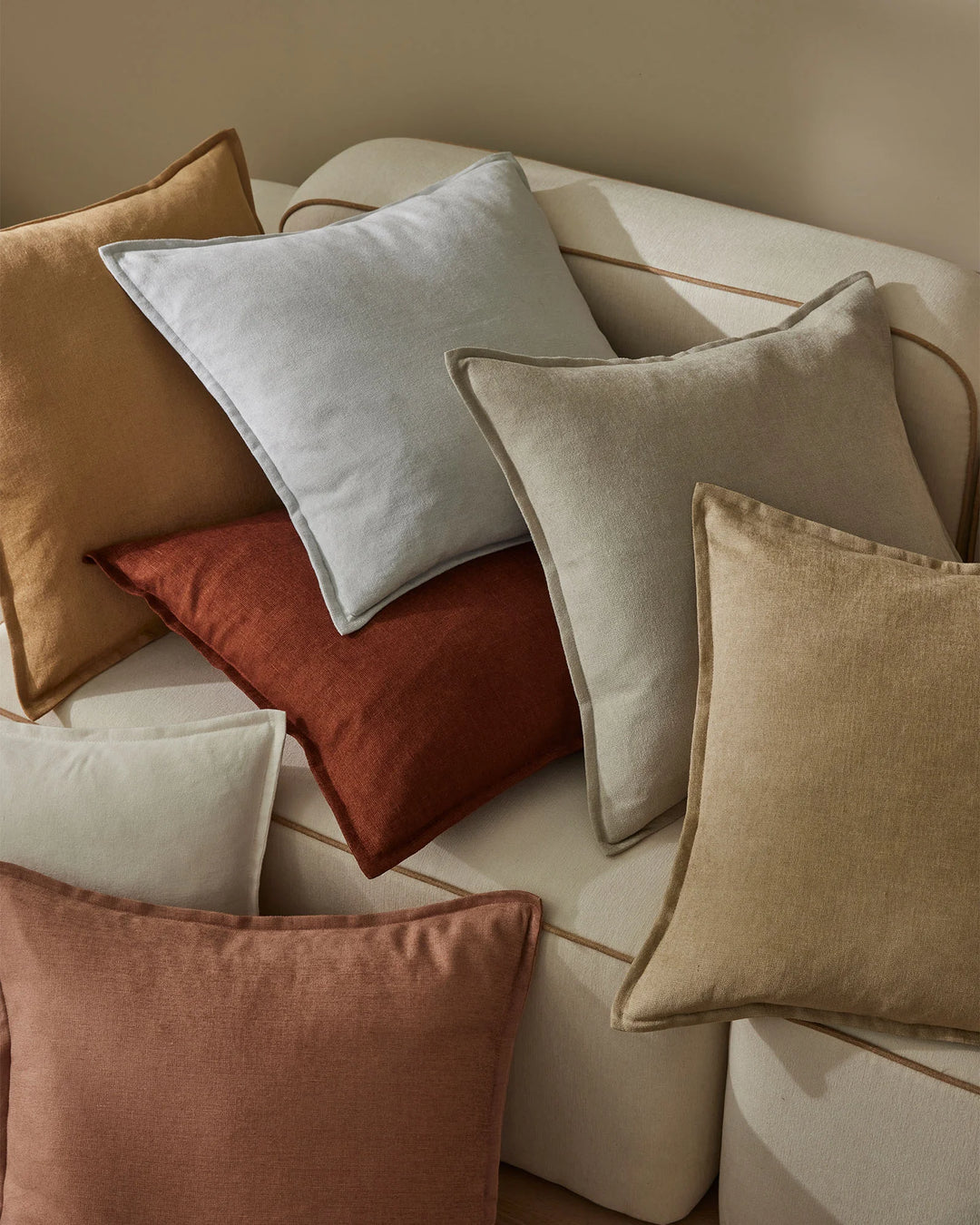 Curated stack of linen viscose cushions in warm terracotta, sage, cream, and soft taupe tones; earthy, neutral palette with textured fabric weaves and piped edges arranged on neutral base for modern interior styling.