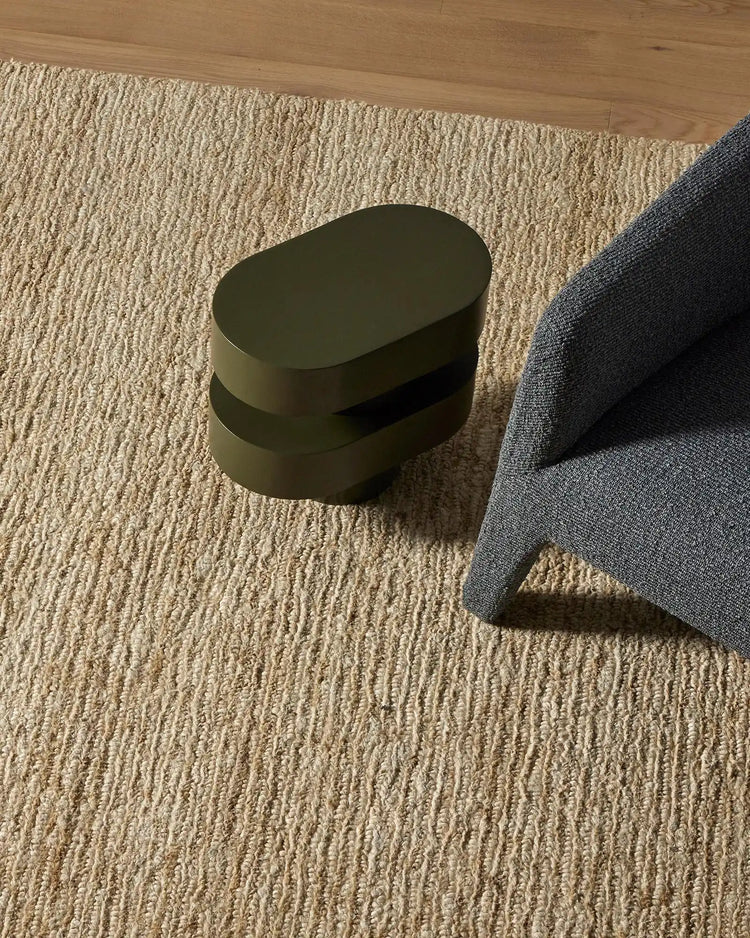 Neutral-toned living space with cream textured Bastian rug, charcoal fabric edge, and olive cylindrical side table casting soft shadows on jute flooring
