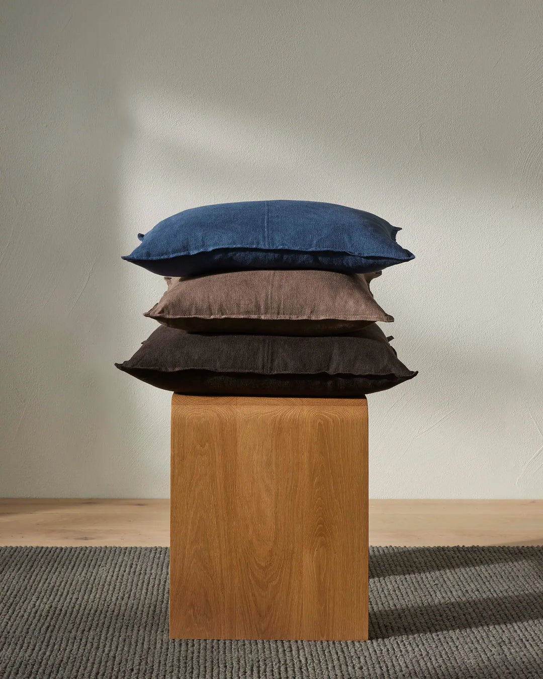 Stacked linen cushions in navy, taupe and charcoal tones on wooden plinth, neutral minimalist styling with woven rug and soft natural light