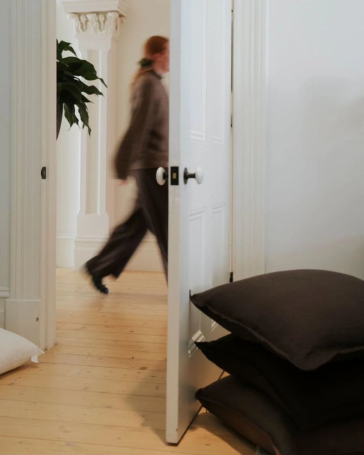 Minimalist hallway with stacked black linen cushions, white panelled doors and natural timber floors, neutral palette with green plant accent