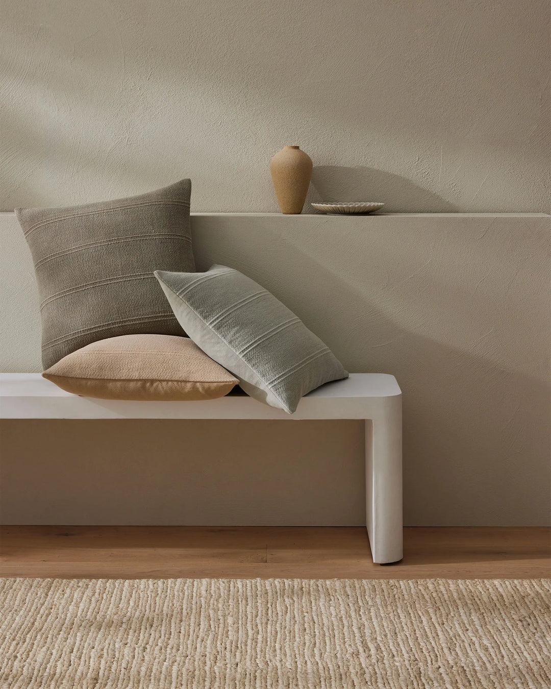 Minimalist living room styled with Palma cushions in neutral tones, featuring striped linen textures, warm wood flooring, and organic ceramic vessels on a platform bench