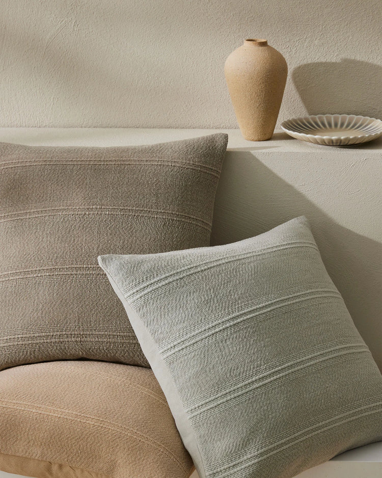 Neutral minimalist living room styling with layered Palma cushions in stone, shell and ice tones, textured linen cotton fabrics, and earthy ceramic accessories on white walls