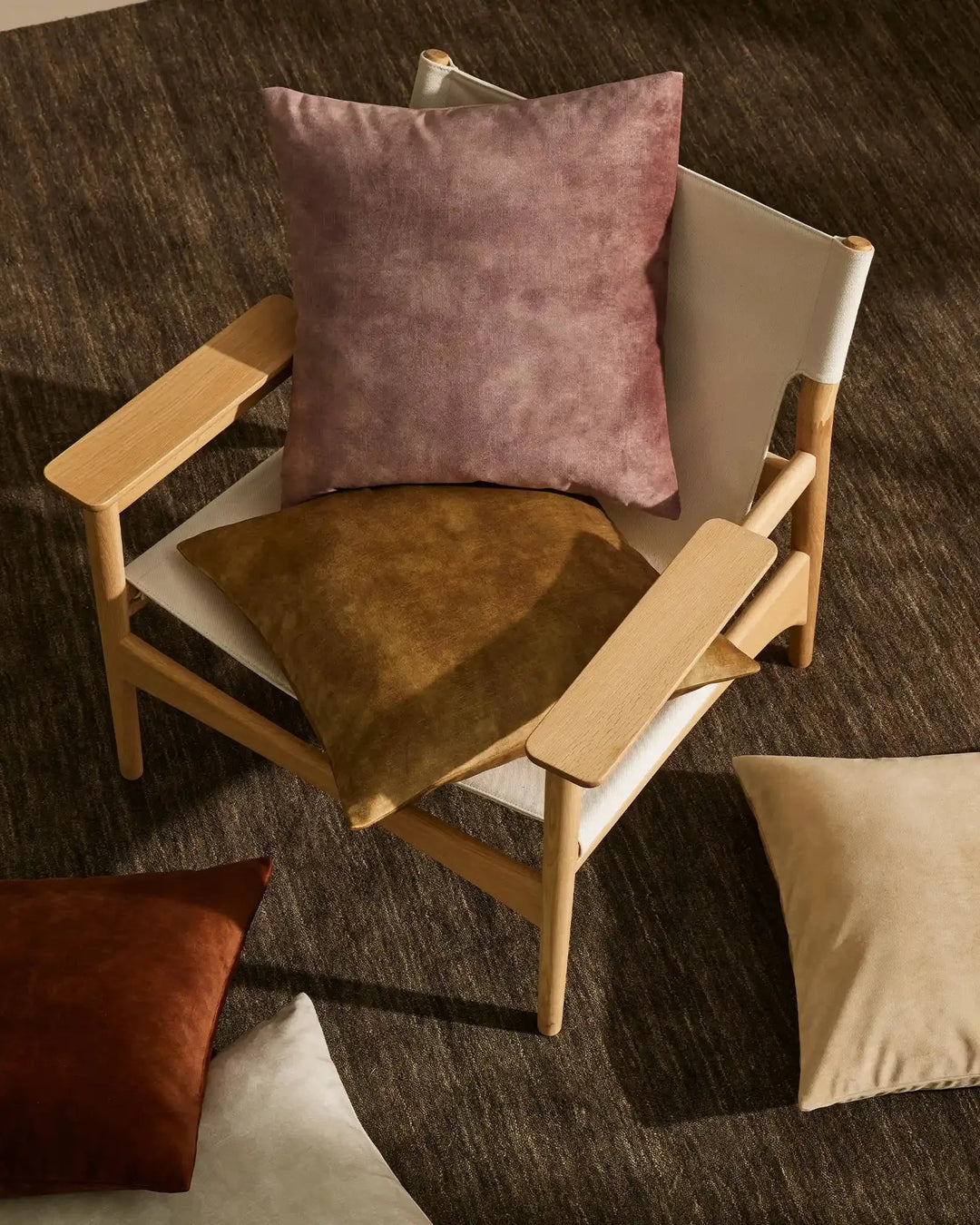 Mid-century wooden armchair styled with mauve velvet and ochre suede cushions on charcoal textured rug, earthy Scandinavian-inspired living room aesthetic