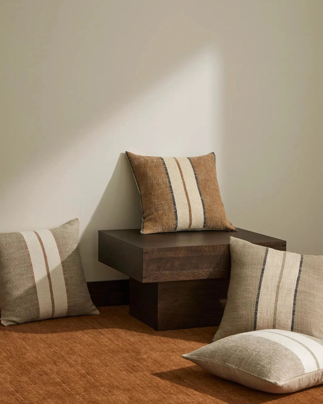 Neutral earthy lifestyle styling with Ottavio cushions in warm terracotta, sage, and taupe tones featuring cream stripe detailing, arranged on dark timber blocks against warm-toned flooring
