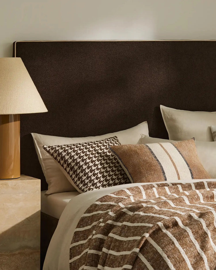 Neutral bedroom styled with chocolate brown headboard, cream pillows, and layered geometric patterned cushions in houndstooth and striped designs. Warm earth-toned bedding with ivory accents creates a modern, textured aesthetic.