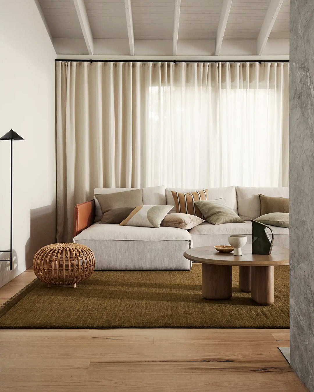 Minimalist living room with neutral palette featuring cream linen sofa, layered beige and sage cushions, olive high-traffic rug, and floor-to-ceiling curtains in soft linen tones
