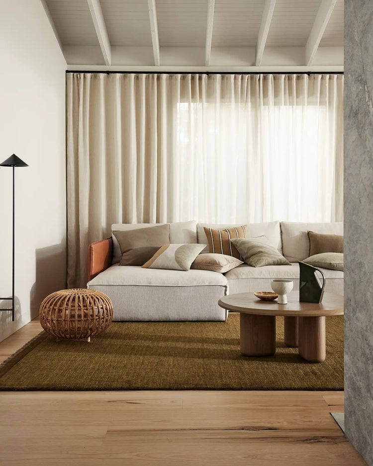 Minimalist living room with neutral palette featuring cream linen sofa, layered beige and sage cushions, olive high-traffic rug, and floor-to-ceiling curtains in soft linen tones