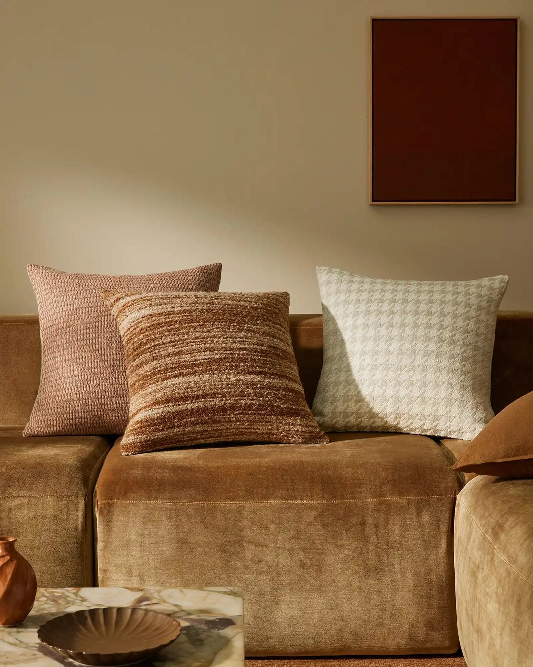 Warm earth-tone living room with layered cushions in blush, caramel and cream on a cognac sofa, featuring textured linen and wool blends against soft beige walls and natural light