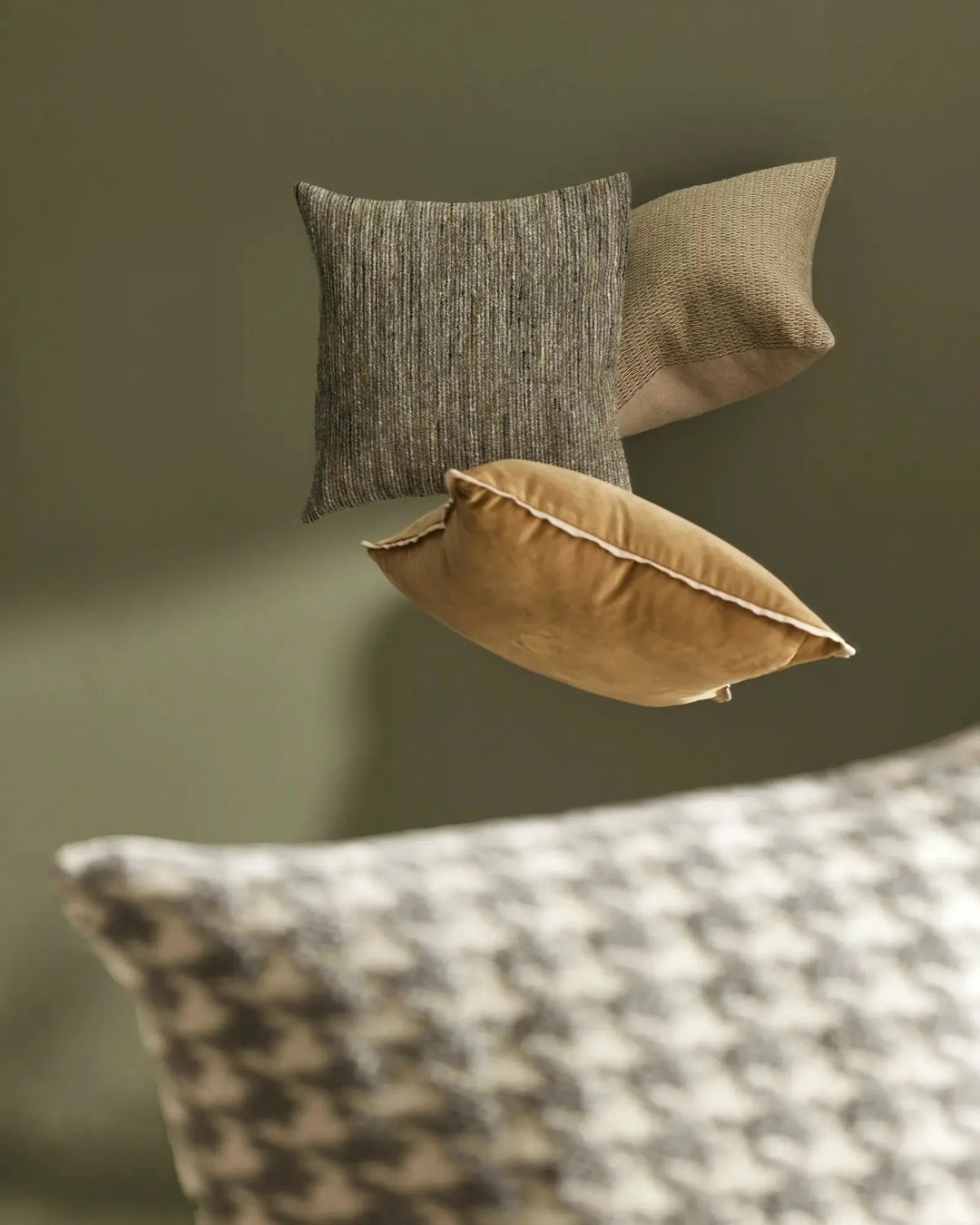 Neutral earth-tone cushion styling with sage green wall backdrop. Textured linen blend pillows in taupe, cream and warm sand tones layered together, showcasing woven surface details and soft fabric drape.