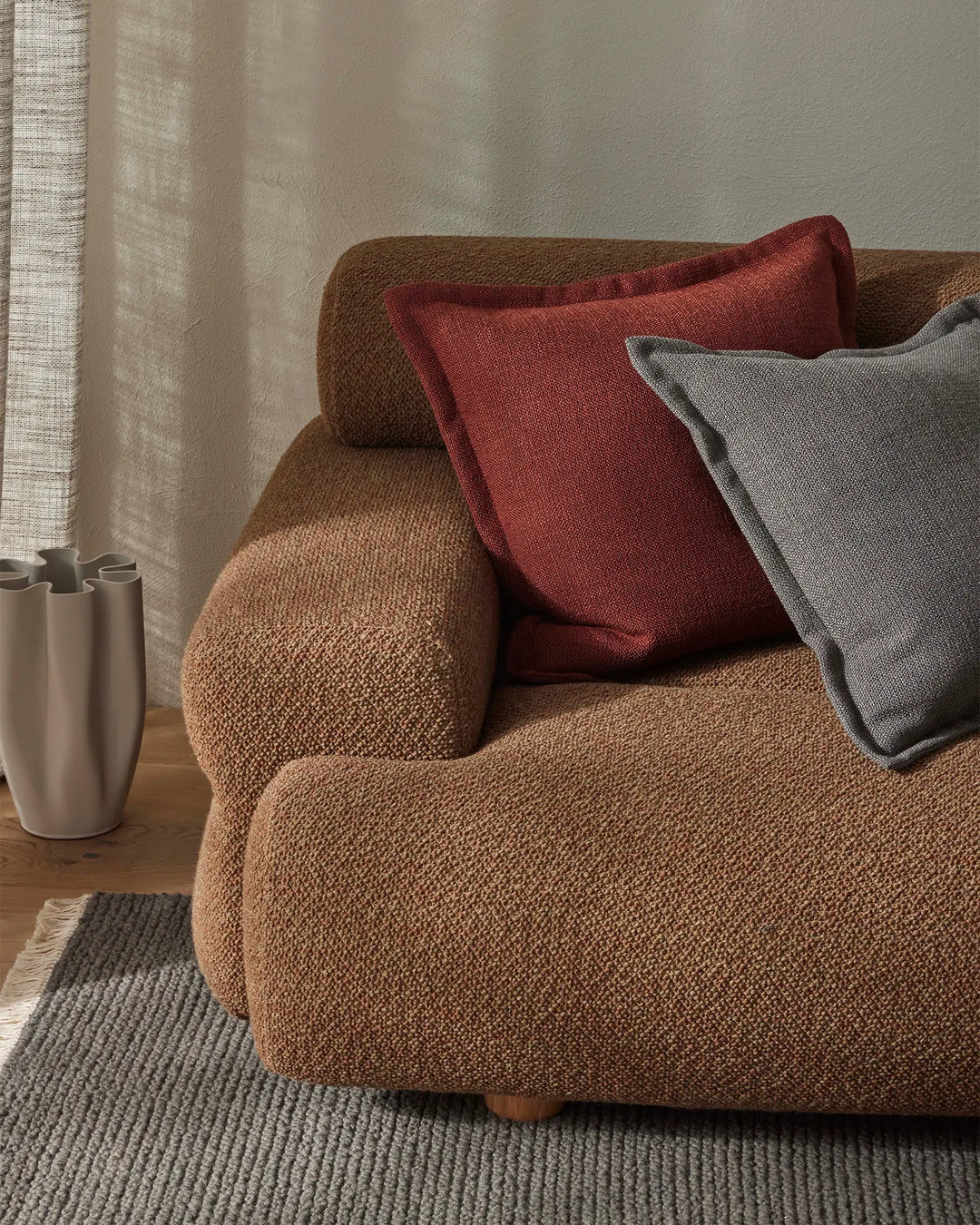 Warm neutral living room with terracotta armchair layered with paprika and graphite cushions, linen curtains, textured rug and ceramic vessel styling