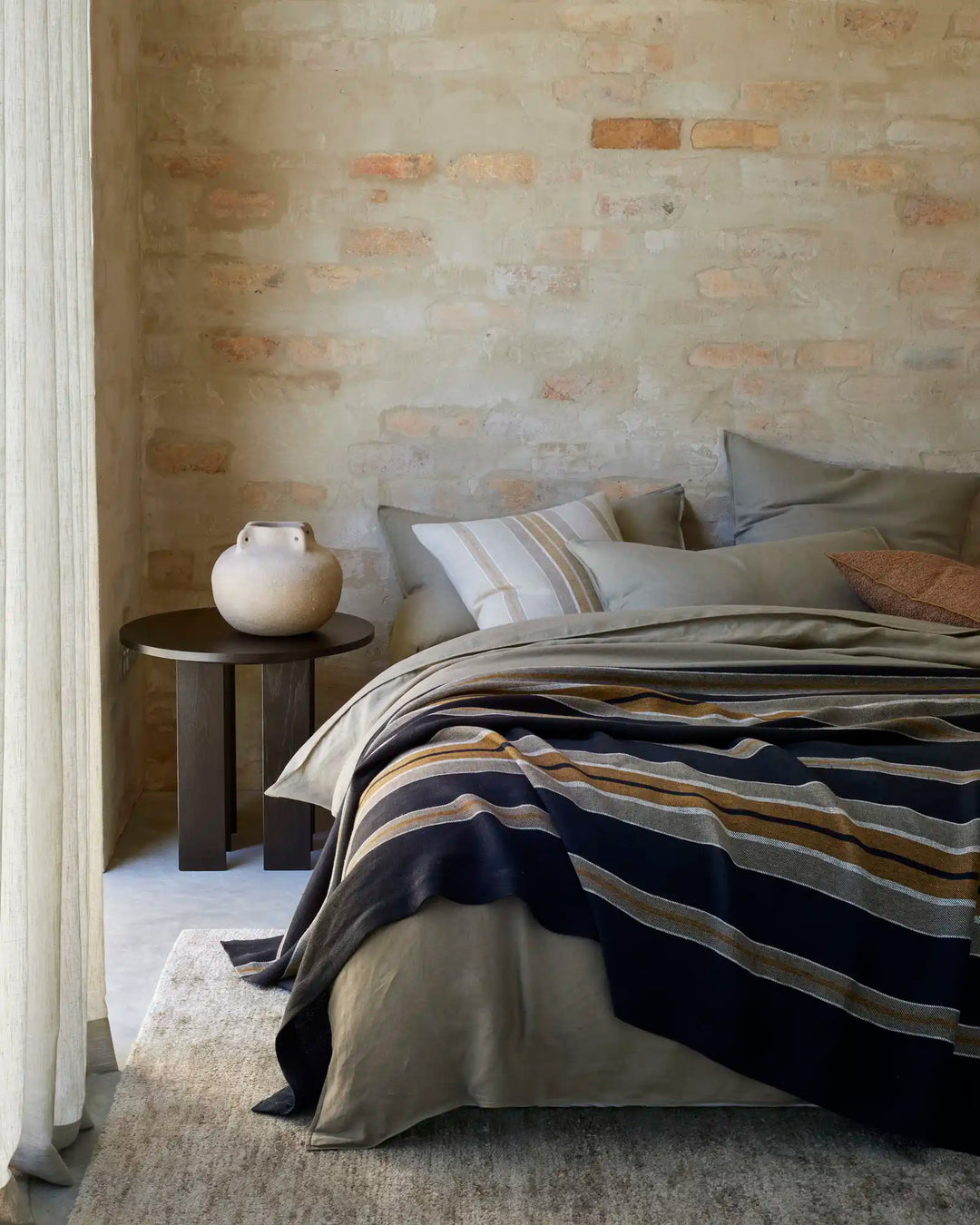 Minimalist bedroom with exposed brick wall, Ravello Quilt Cover and Fitted Sheet in caper tones layered with navy striped bedding, natural linen textures, and earthy neutral palette