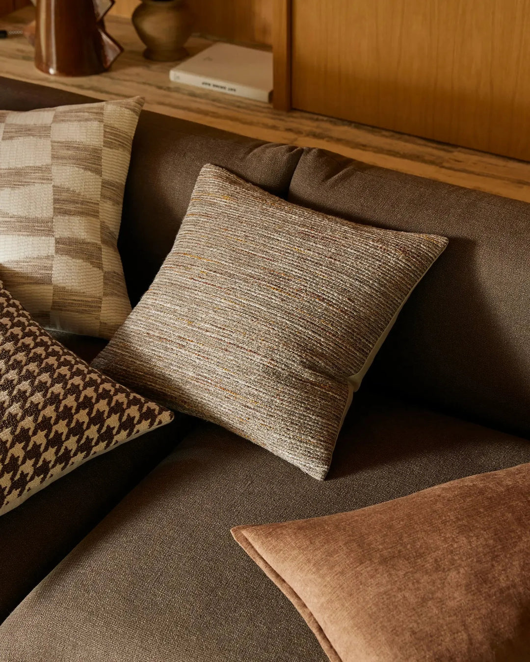 Warm neutral living room styled with layered textured cushions in cream, taupe and toffee tones on a sage sofa, featuring Vista Natural and Fiore ranges with woven patterns and linen blends