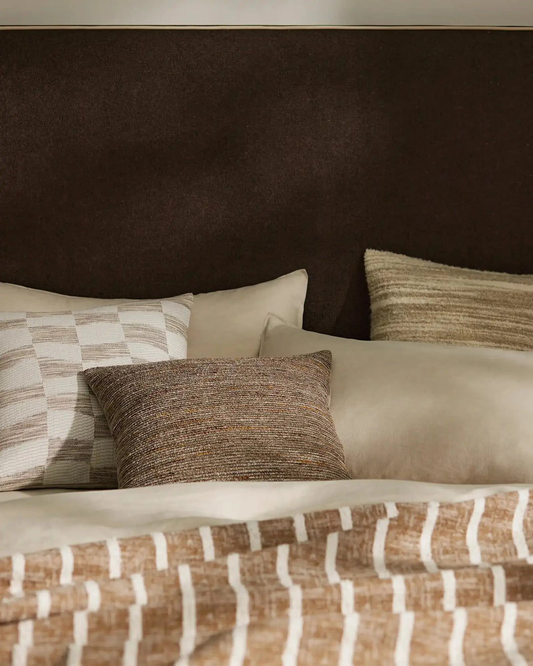 Neutral bed styling with layered textures: woven cushions in taupe and natural tones, cream linens, and a geometric patterned throw against a charcoal headboard