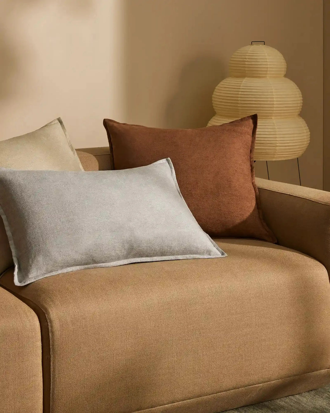 Warm earthy living room with layered Fiore cushions in toffee, sage and breeze tones on a camel sofa, paired with a woven paper pendant light