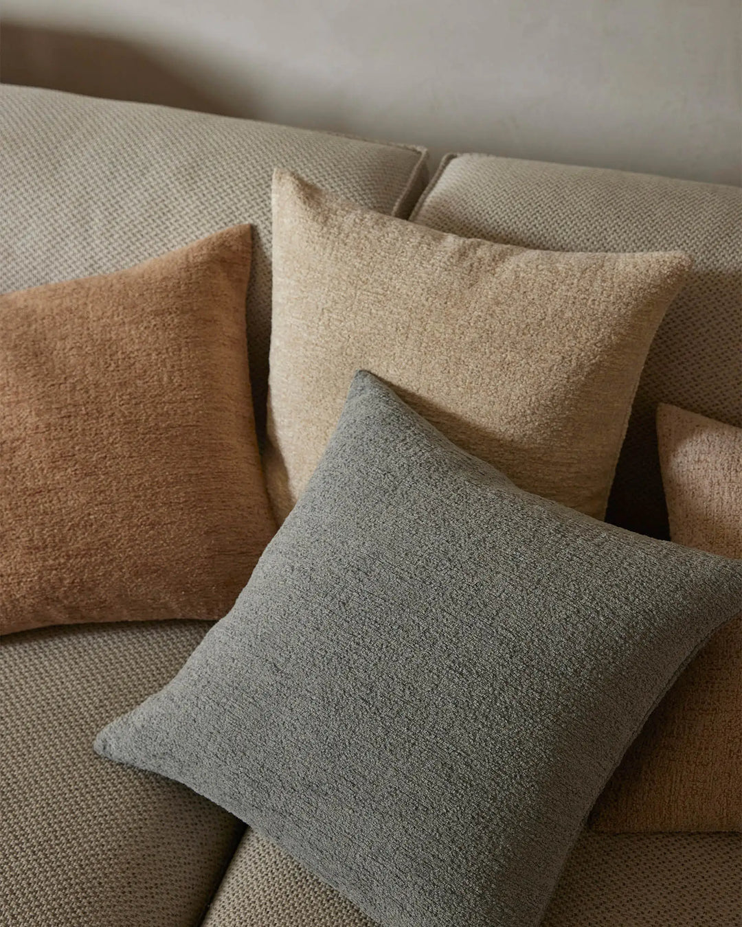 Neutral cushions in warm caramel, cream and grey tones layered on a pale linen sofa, showcasing textured fabrics and earthy, minimalist styling